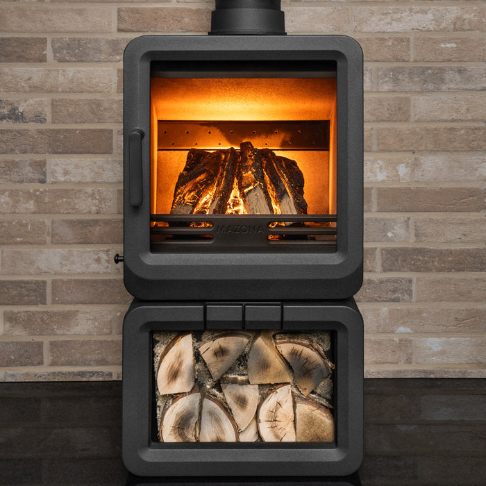 A black wood-burning stove with a lit fire inside and a lower compartment filled with stacked firewood, set against a light brick wall.